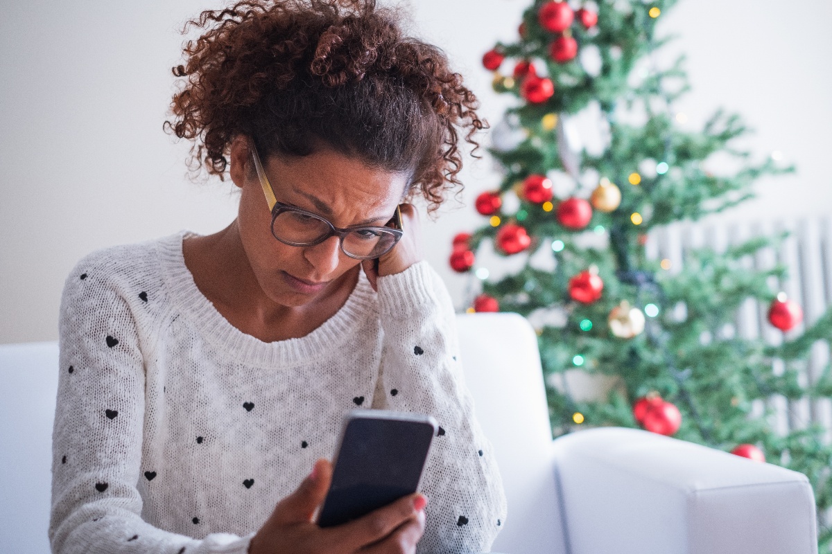 A lonely woman sits in her living room next to a Christmas tree while using her cell phone to access the Pyx Health app.