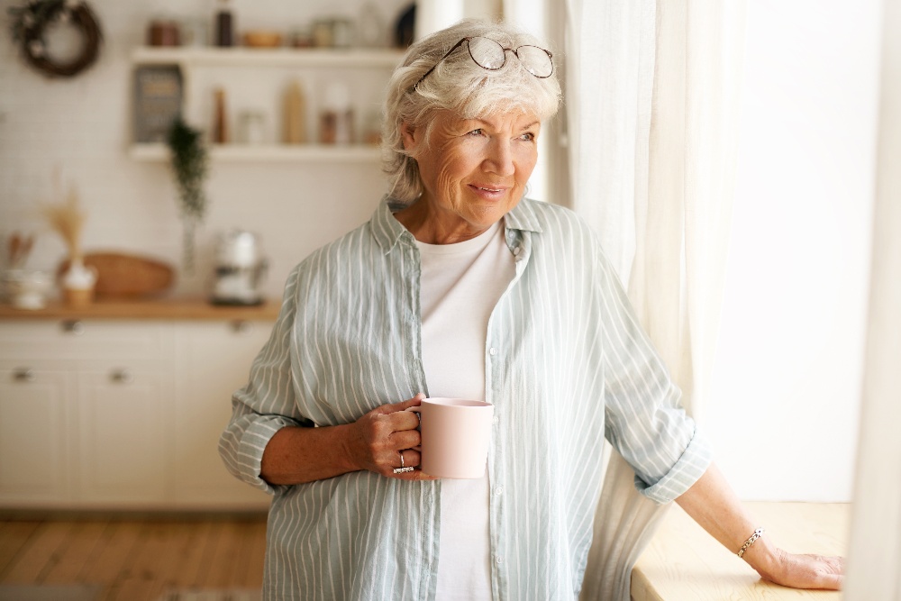 An older woman holds a cup of coffee while looking out a window.