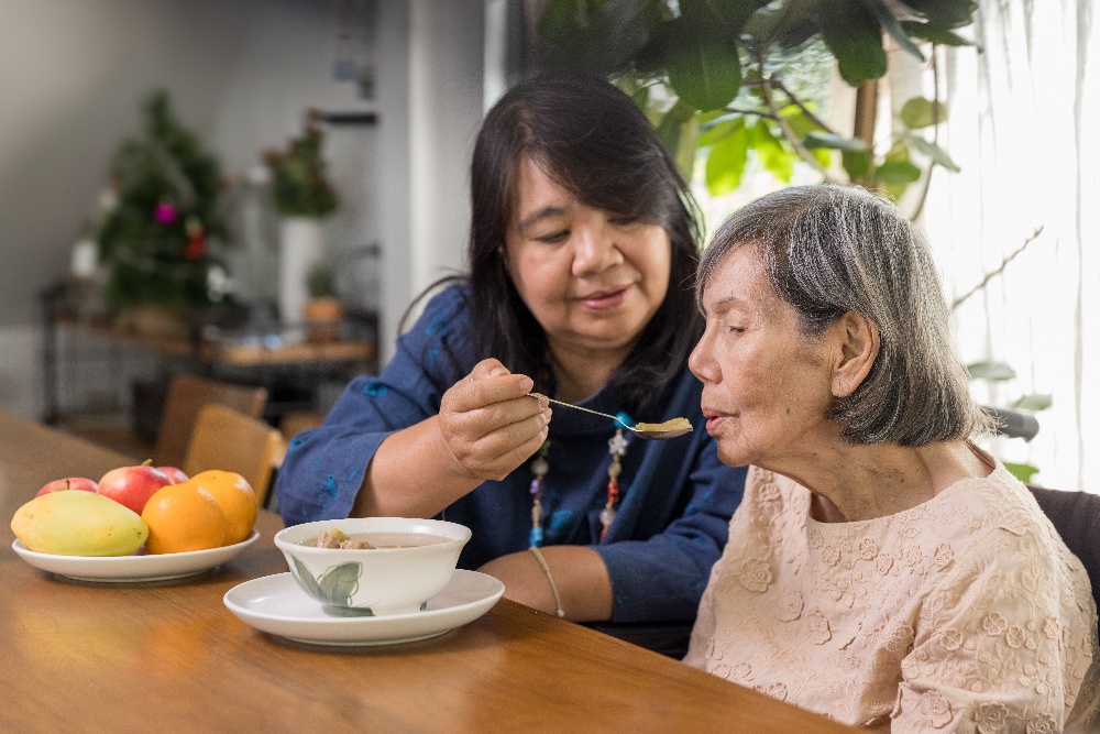 A middle-aged female caregiver feeds soup to an elderly patient with a spoon. 