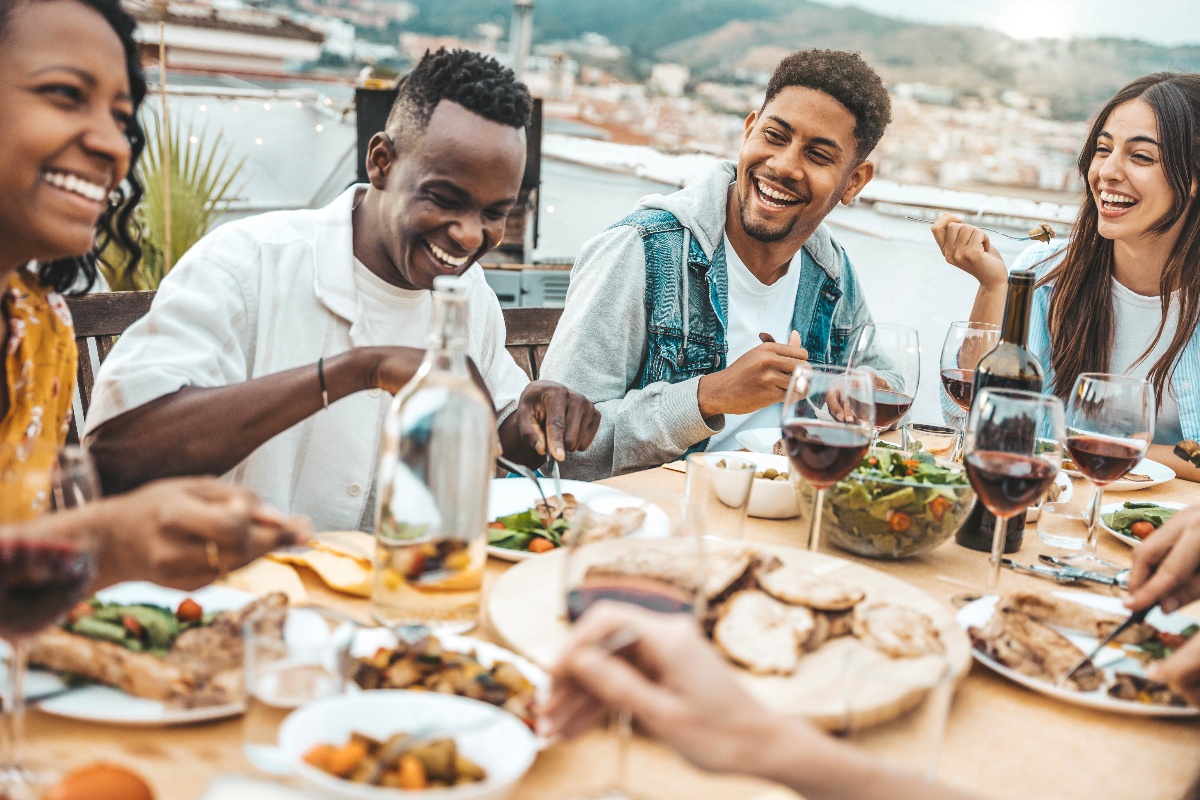 A diverse group of young adults laughing while enjoying a rooftop dinner together. 