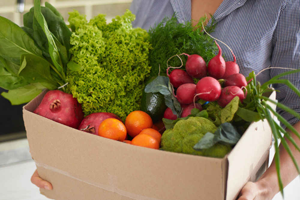 A woman carries a cardboard box filled with groceries from a food delivery.