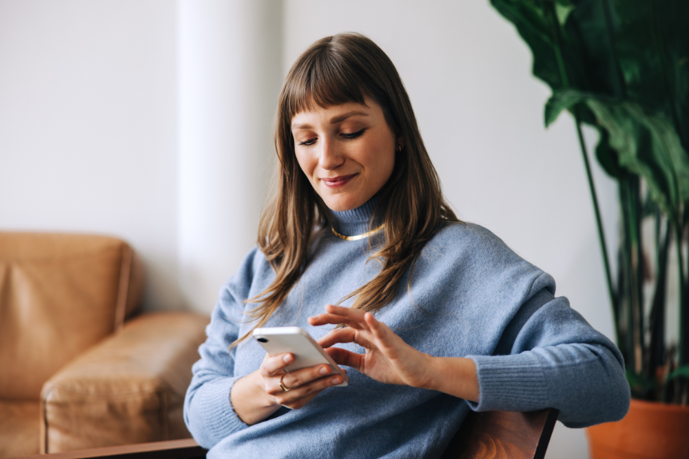 A woman uses her cellphone to access the Pyx Health app while sitting on a sofa. 