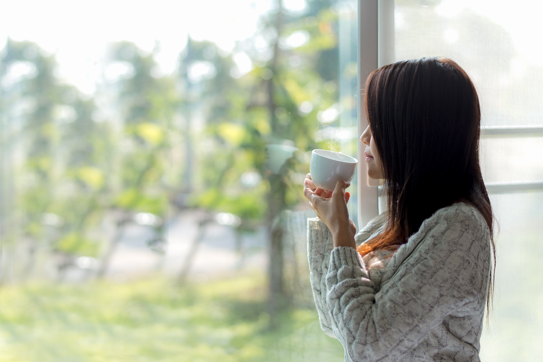 A woman holds a cup of coffee as she stares out of a window.