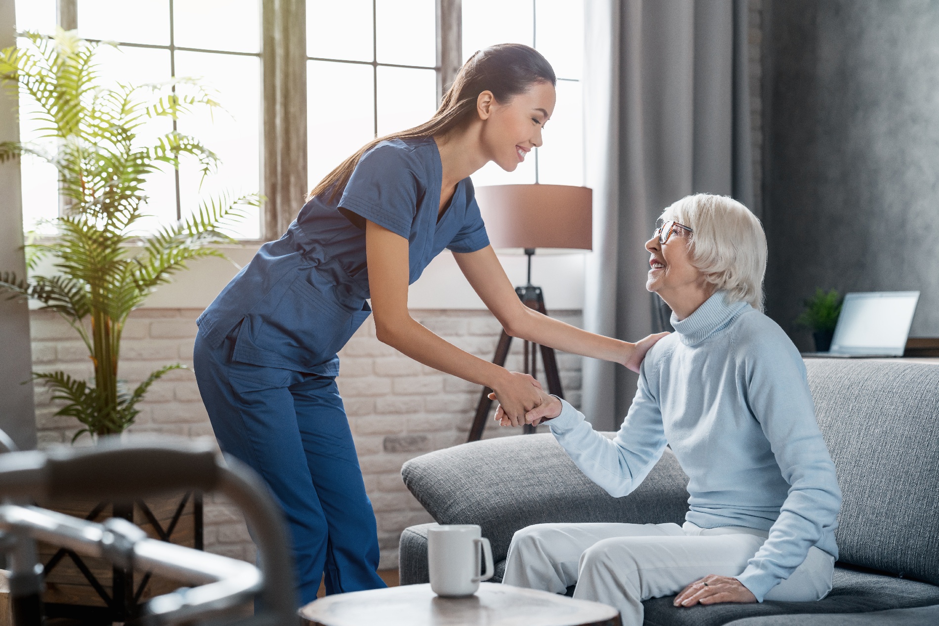 An older woman is being helped up off a sofa by a female nurse.