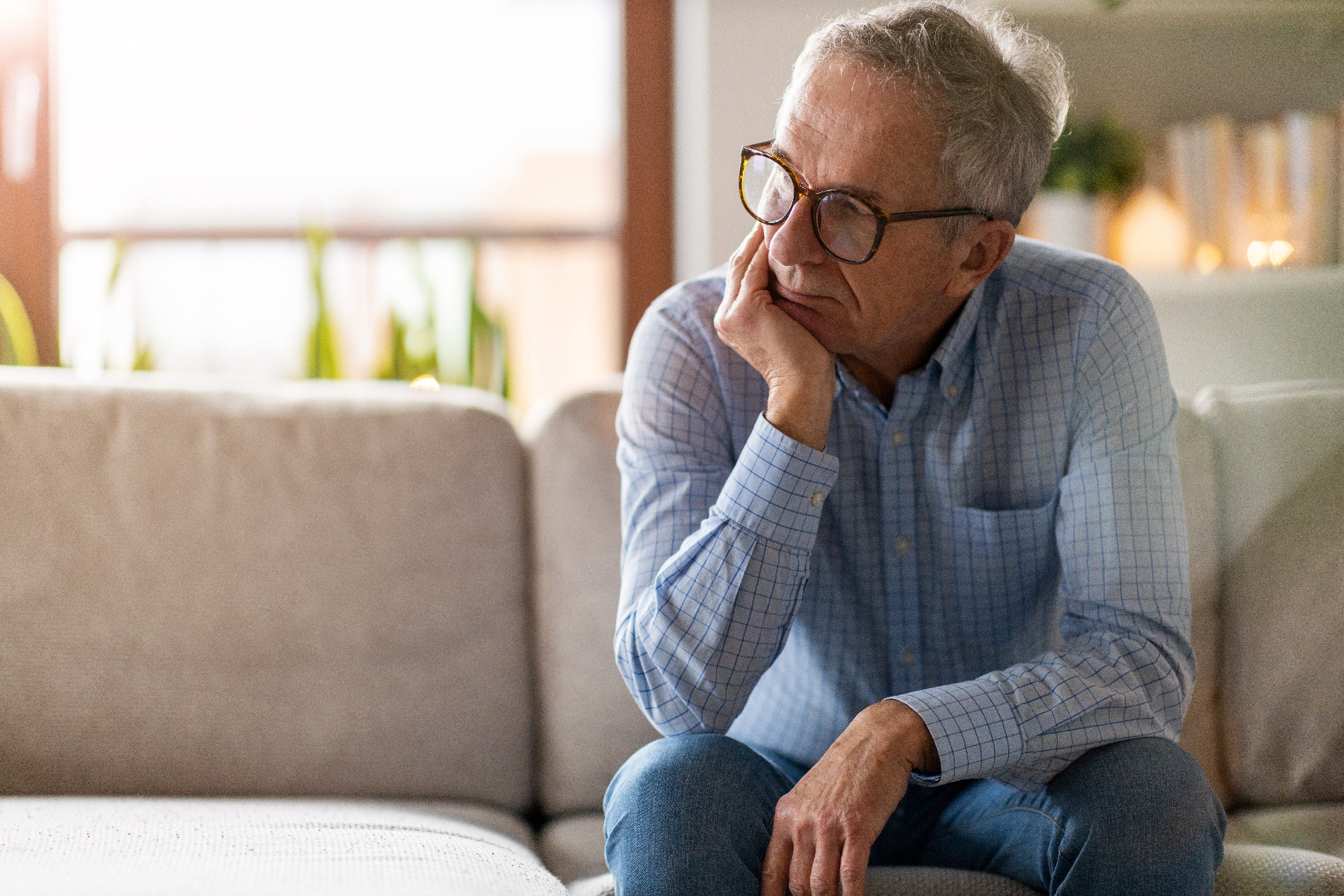 An older man holds his face as he thinks while sitting on a sofa.