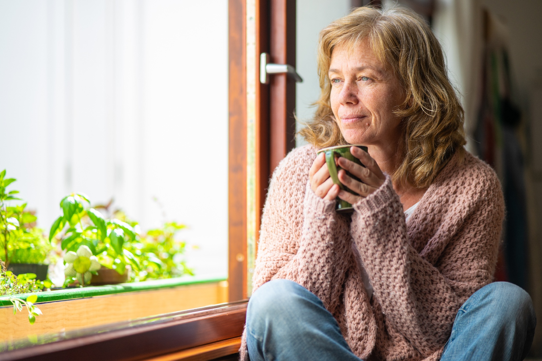 An older woman holds a mug with both hands while looking out a window lined with houseplants. 
