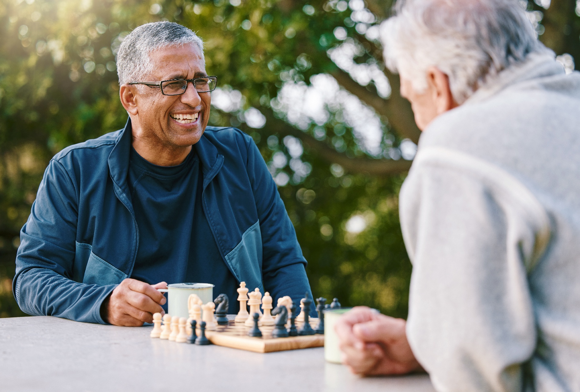 Two older men play a game of chess and drink coffee while sitting in a beautiful park.