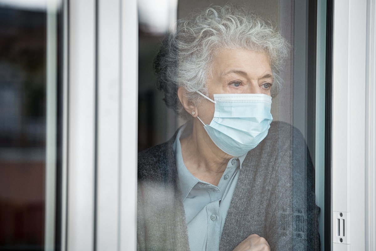 An elderly woman looks out a window at home while wearing a mask to protect against COVID-19.